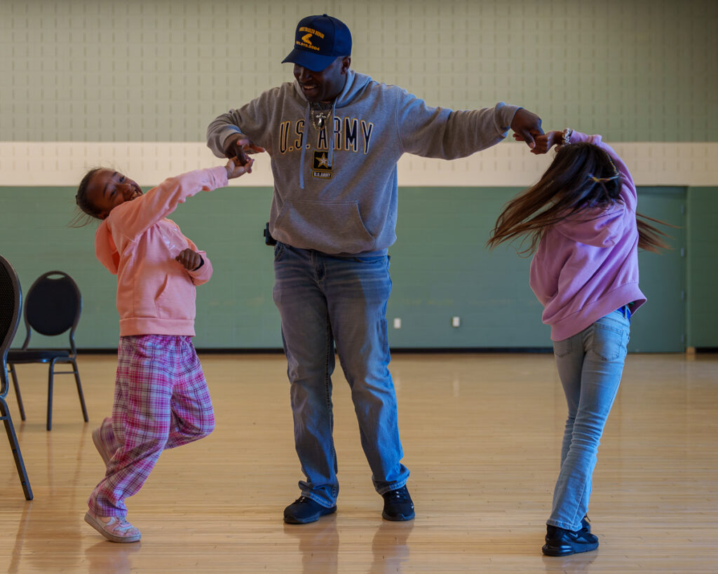 A man dances joyfully with two young girls in a sunlit gymnasium during a music event.