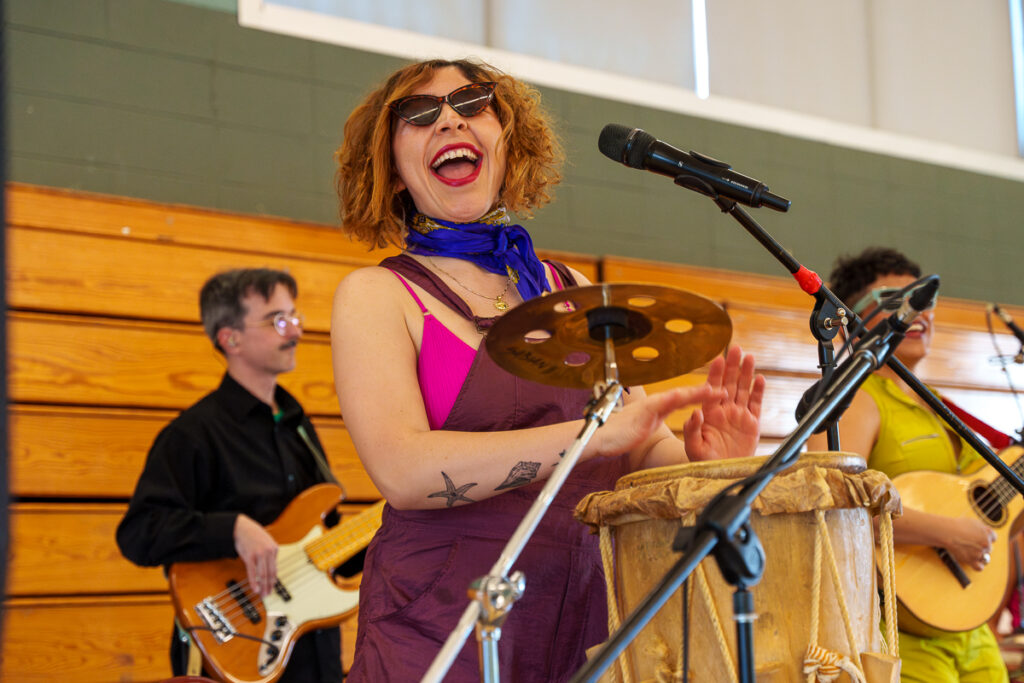 A smiling musician claps along behind a drum and microphone, wearing bright clothing and sunglasses, while fellow bandmates play nearby