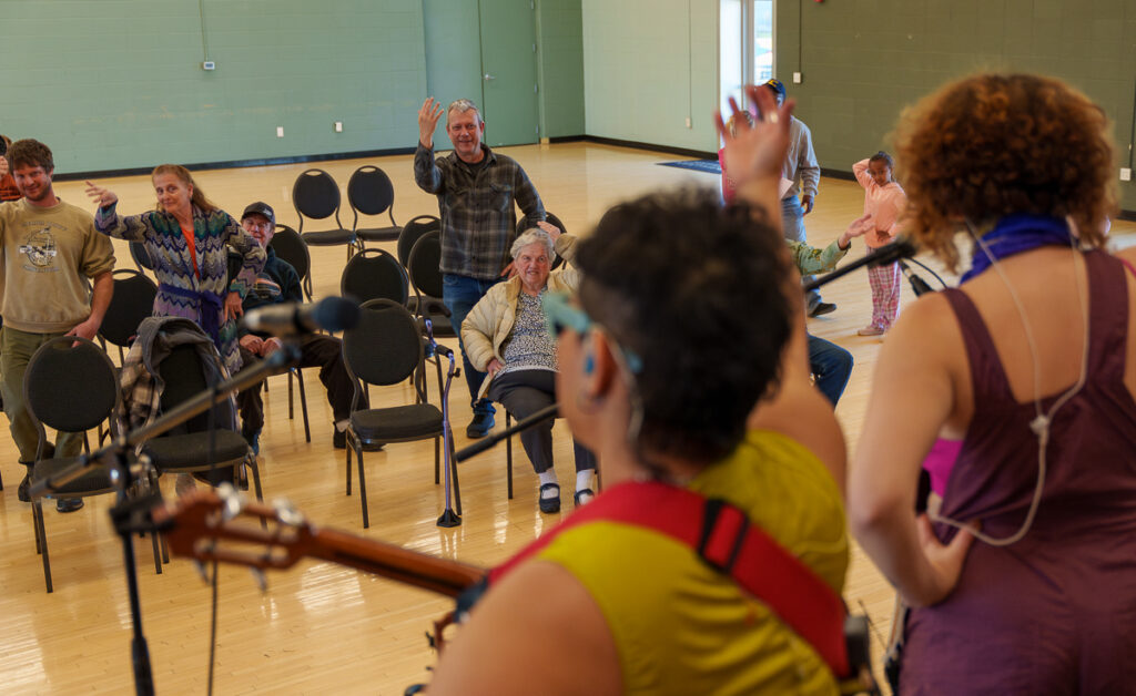 Audience members of all ages smile and wave from their seats in a community center as musicians perform just out of frame.
