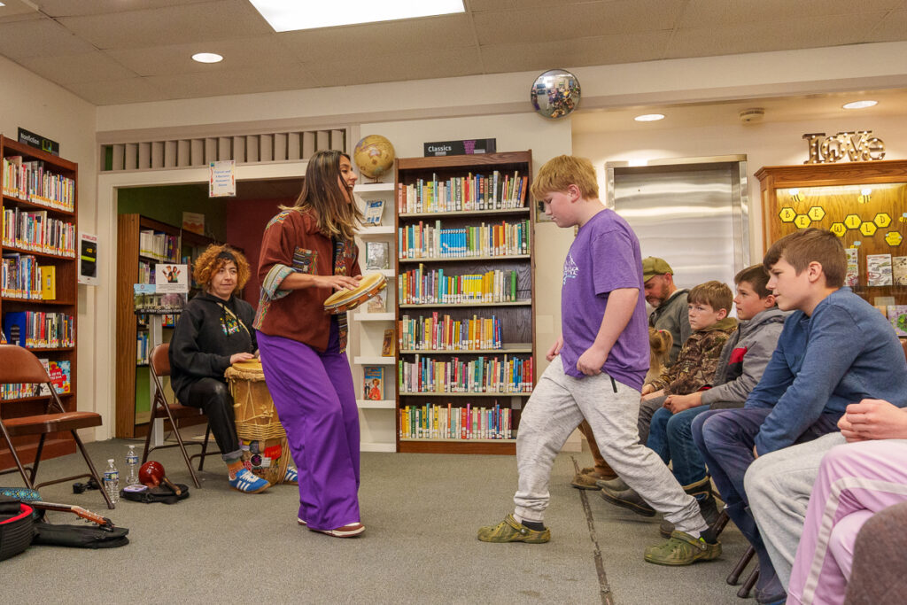 A woman holding percussion instruments engages with a boy in a purple shirt during a musical workshop at a library.