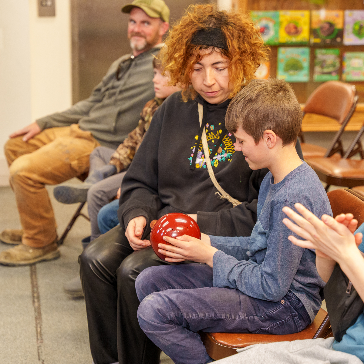 A seated musician assists a young boy as he explores a round percussion instrument, with other attendees watching nearby.