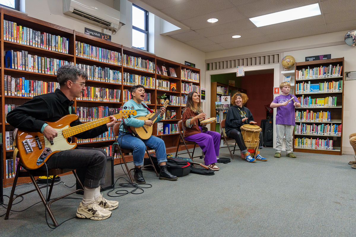 A band performs an interactive music program in a library, joined by an enthusiastic young participant standing at the front.