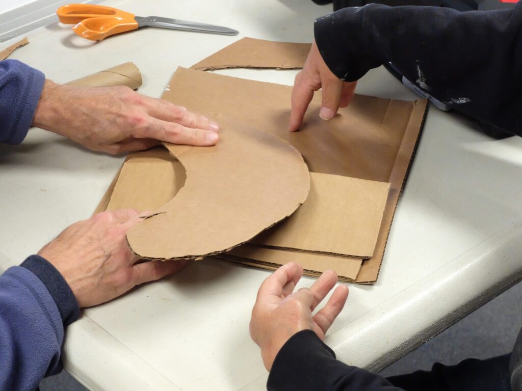 Two sets of light-skinned hands touch and adjust pieces of brown cardboard on a white table.