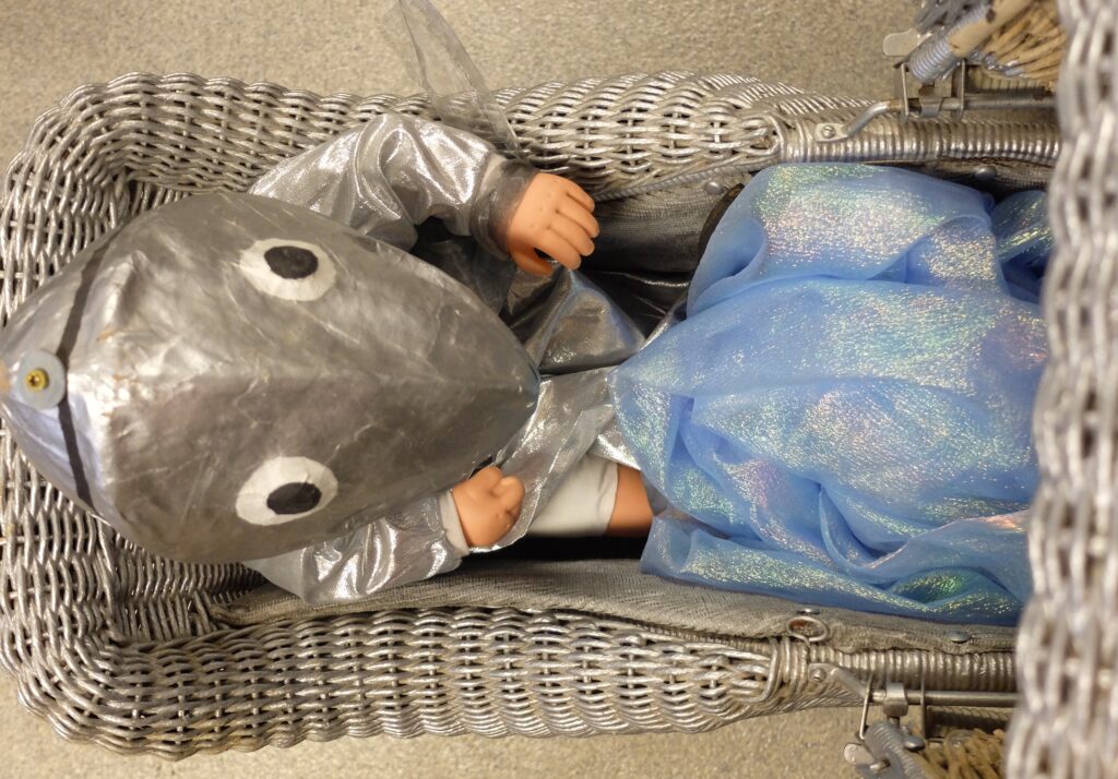 A silver baby carriage holds a silver fish head over a doll toy.