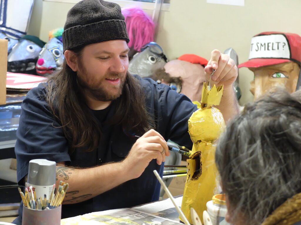 A light-skinned person wearing a hat paints gold cardboard.