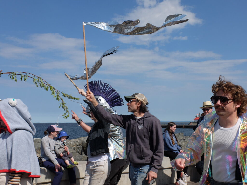 People hold up silvery fish flags against a blue sky and lake.
