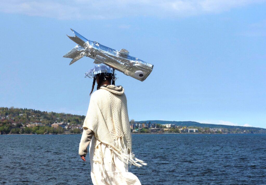 A person wearing all white holds a silver fish puppet in the sky in front of a lake horizon.