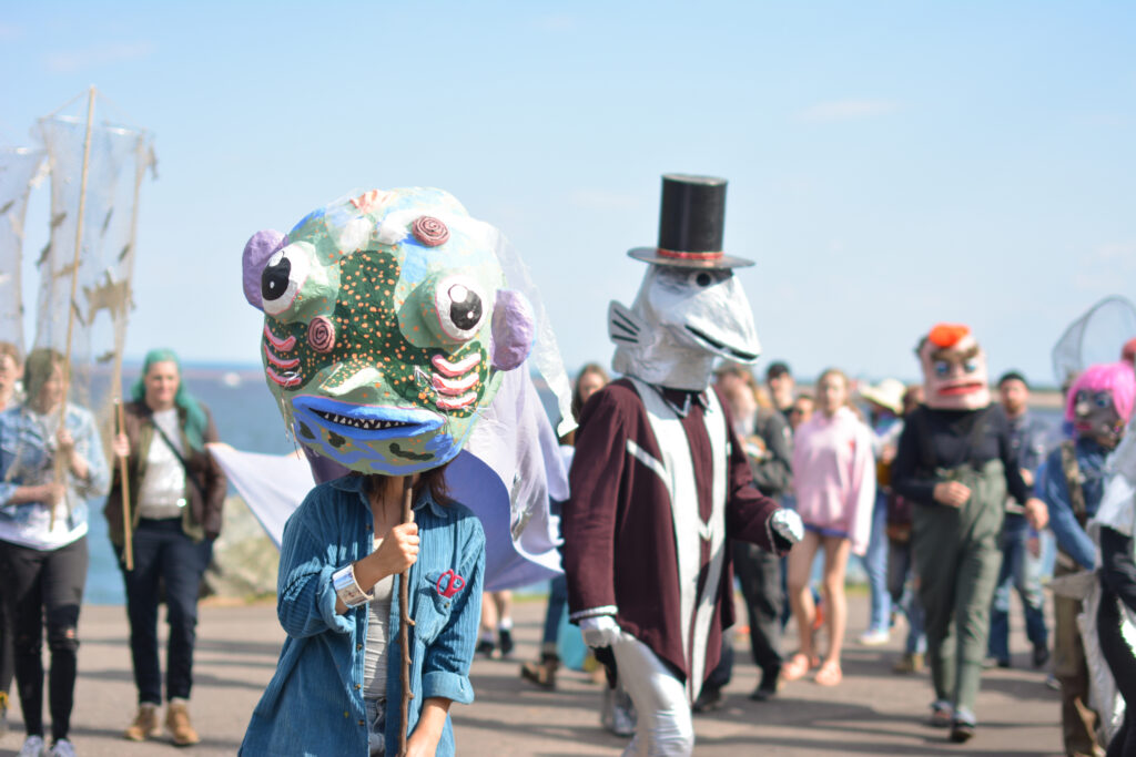 A group of people parade under a blue sky, some wearing giant puppet heads.