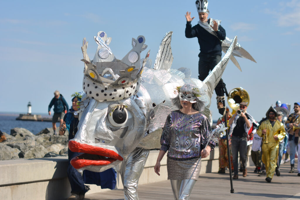 People, including one on stilts, and a large silver fish puppet with red lips wearing a crown, walk down a sidewalk with a lake and lighthouse in the distance.