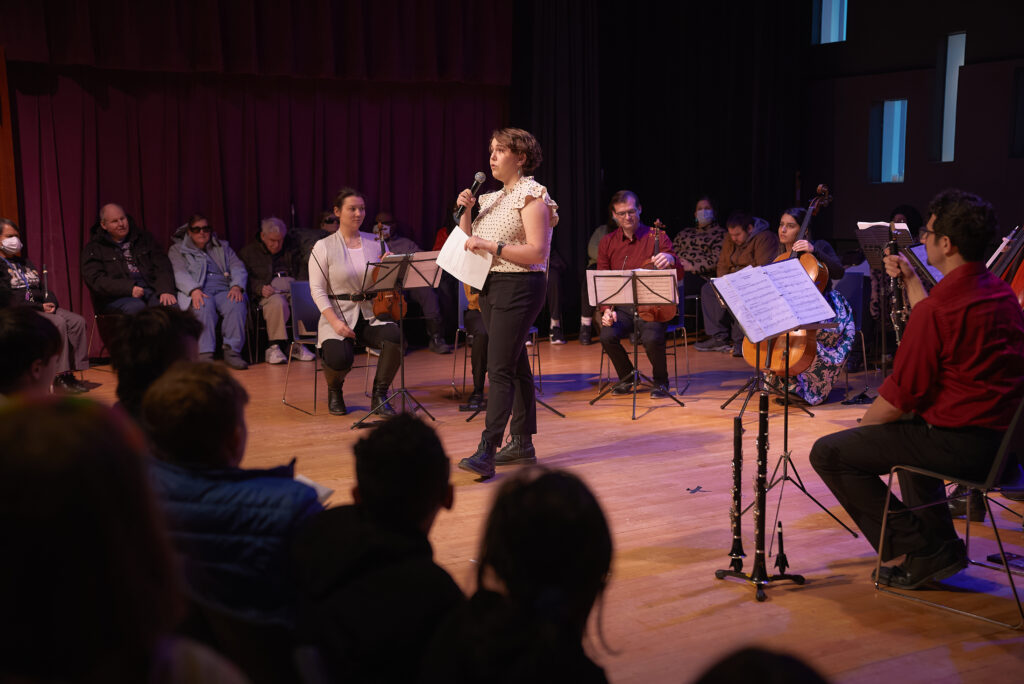 Woman speaking at a microphone onstage surrounded by musicians