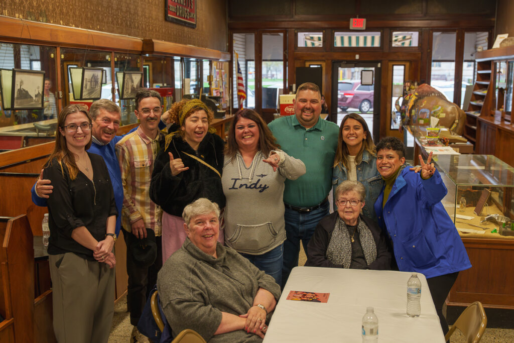 A group of community members and musicians pose together, smiling and making playful hand gestures in a warmly lit diner-style setting.