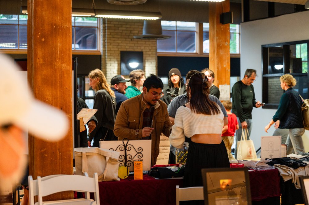 People gather around tables with art for sale inside a building with wooden columns.