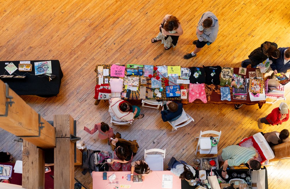 A birds-eye view of vendors sitting at a table on hardwood floors while people stand hearby.