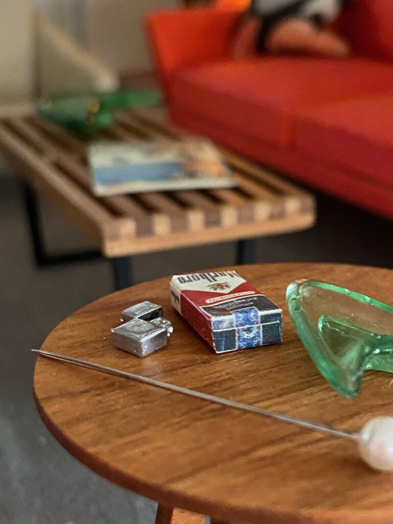 A close up of miniature cigarette box and lighter on a table. Next to it a push pin to show the scale of how tiny the objects are