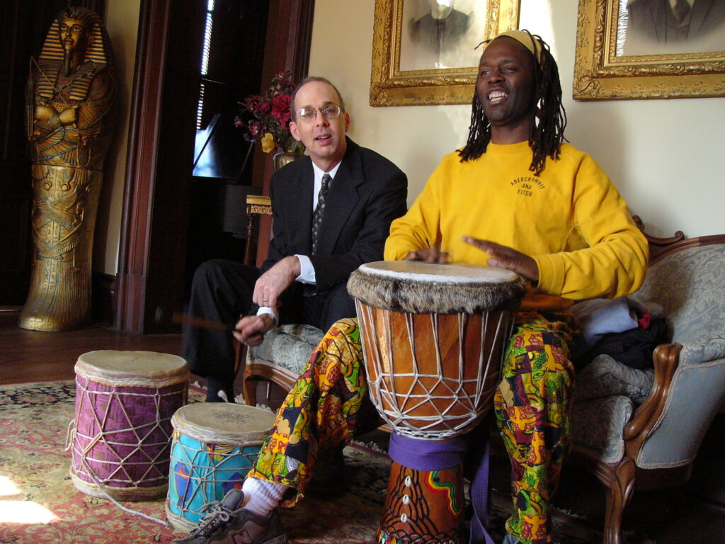 Two men sit indoors surrounded by traditional West African drums. One man in bright patterned pants plays a djembe with joy while the other watches and taps along with a stick.