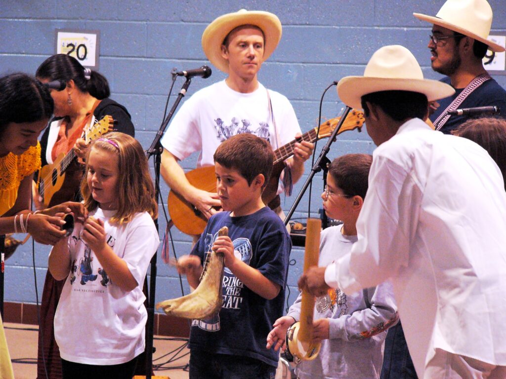 Musicians from Los Utrera stand and play traditional Mexican instruments while children join in, learning to play percussion in a hands-on music activity