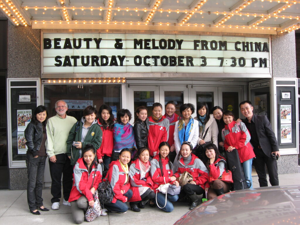 A group of performers and community members pose smiling outside a theater marquee that reads “Beauty & Melody from China – Saturday October 3 7:30 PM.”