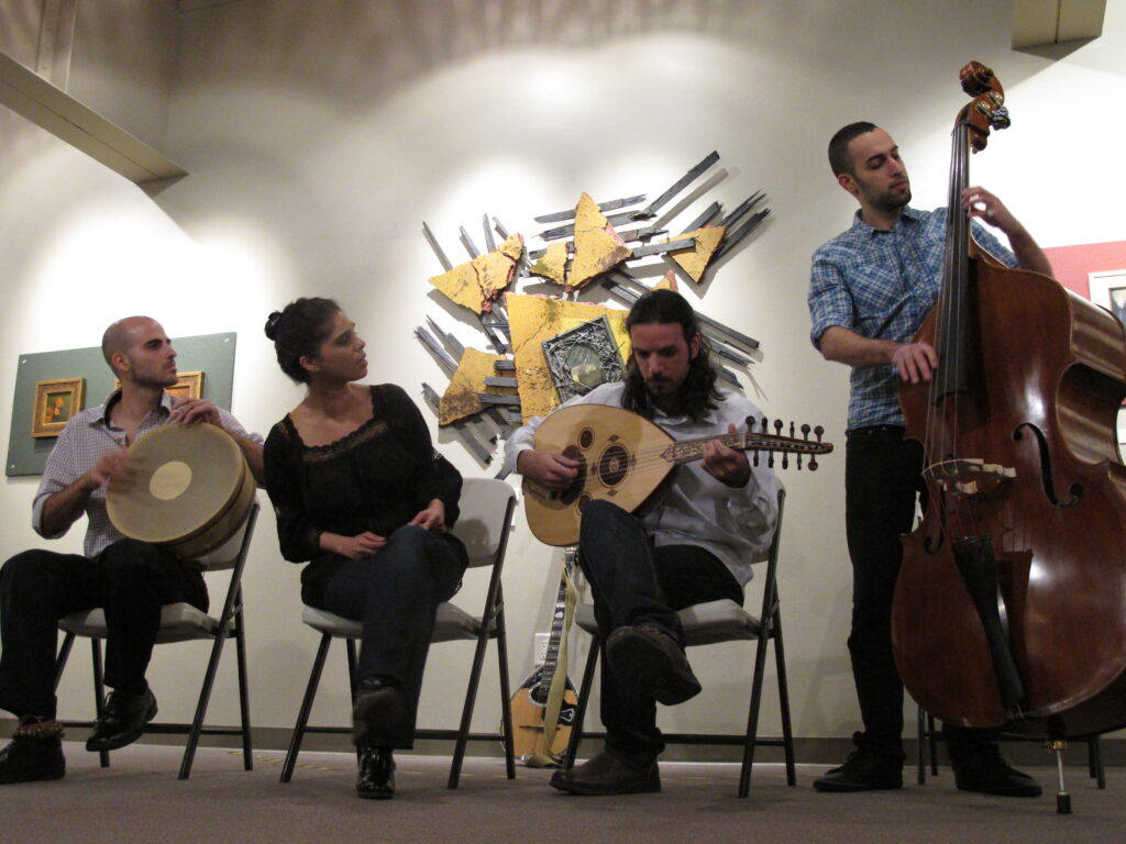 Four musicians sit in a semicircle, playing traditional Middle Eastern instruments, including frame drum, oud, and upright bass, against a backdrop of abstract art