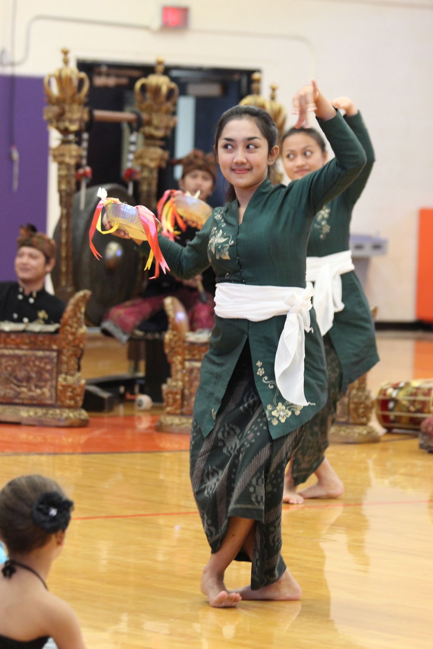 A Balinese dancer in a green outfit with a white sash performs a traditional movement with grace and expression, while seated children watch in awe.