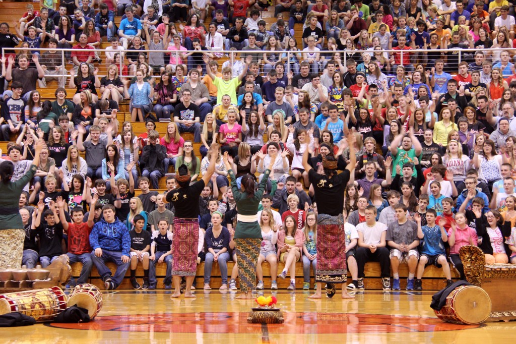 Traditional Balinese performers in green and gold attire lead a crowd of students in an interactive assembly in a packed high school gymnasium.