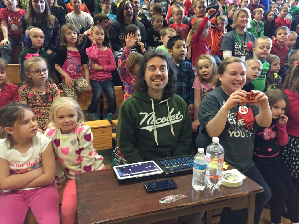 A joyful school audience cheers and claps along with a seated musician and an adult taking photos in the front row of a gymnasium.