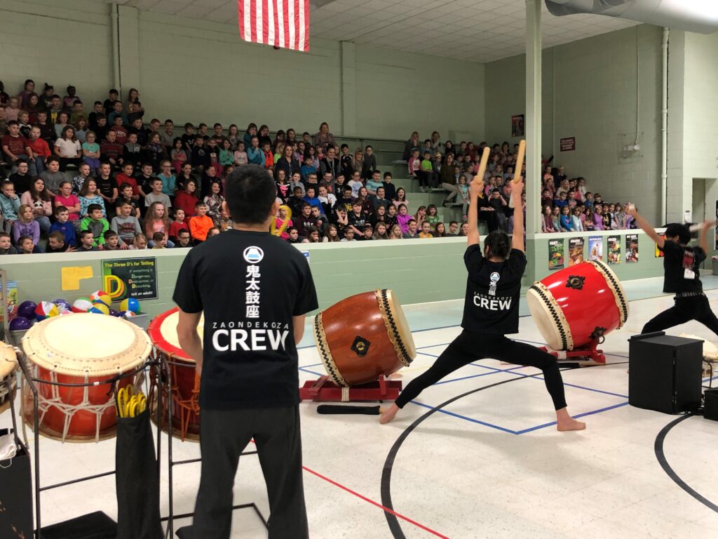 Members of the Ondekoza drumming crew perform with dynamic energy in a gymnasium, while a large group of children watches intently from the bleachers.
