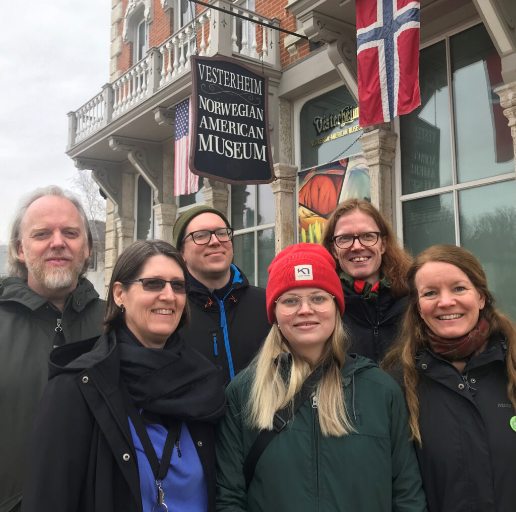 A group of six people poses outdoors in front of the Vesterheim Norwegian-American Museum, smiling in front of an American and Norwegian flag.