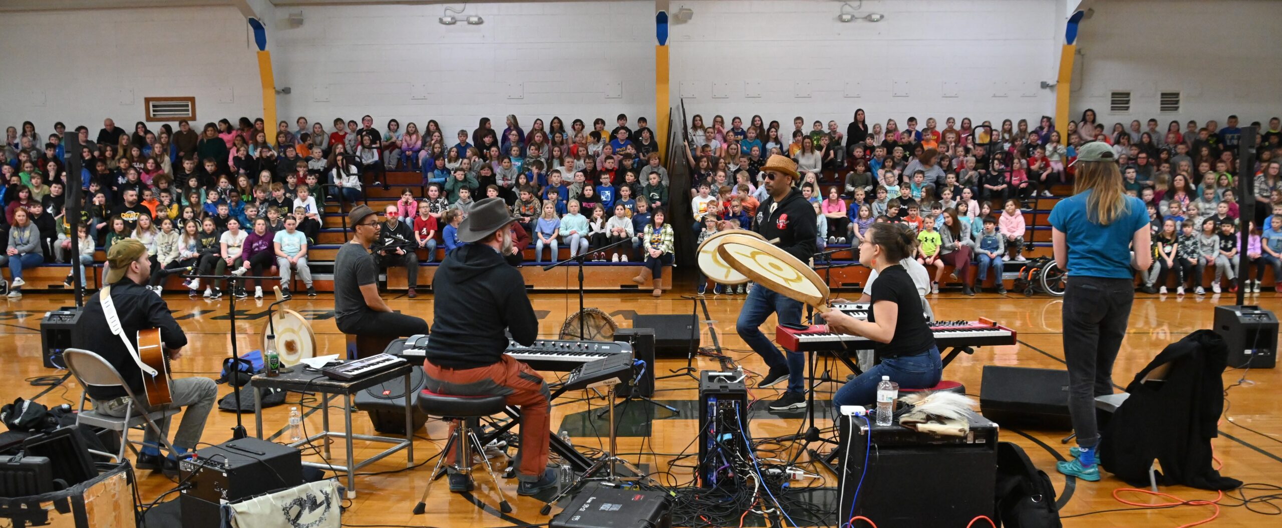 The band Pamyua performs in a school gymnasium surrounded by instruments and sound equipment, as a large audience of children watches from the bleachers.