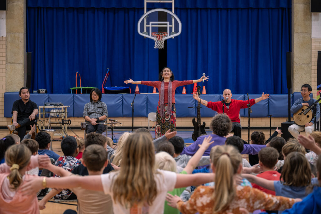 A musician with open arms stands at the center of a school gymnasium stage, beaming at a group of enthusiastic students mirroring the movement