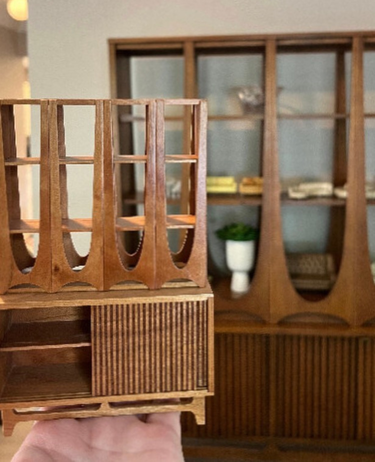 A hand holding up a tiny wooden cabinet furniture with a life-size one of it in view in the background