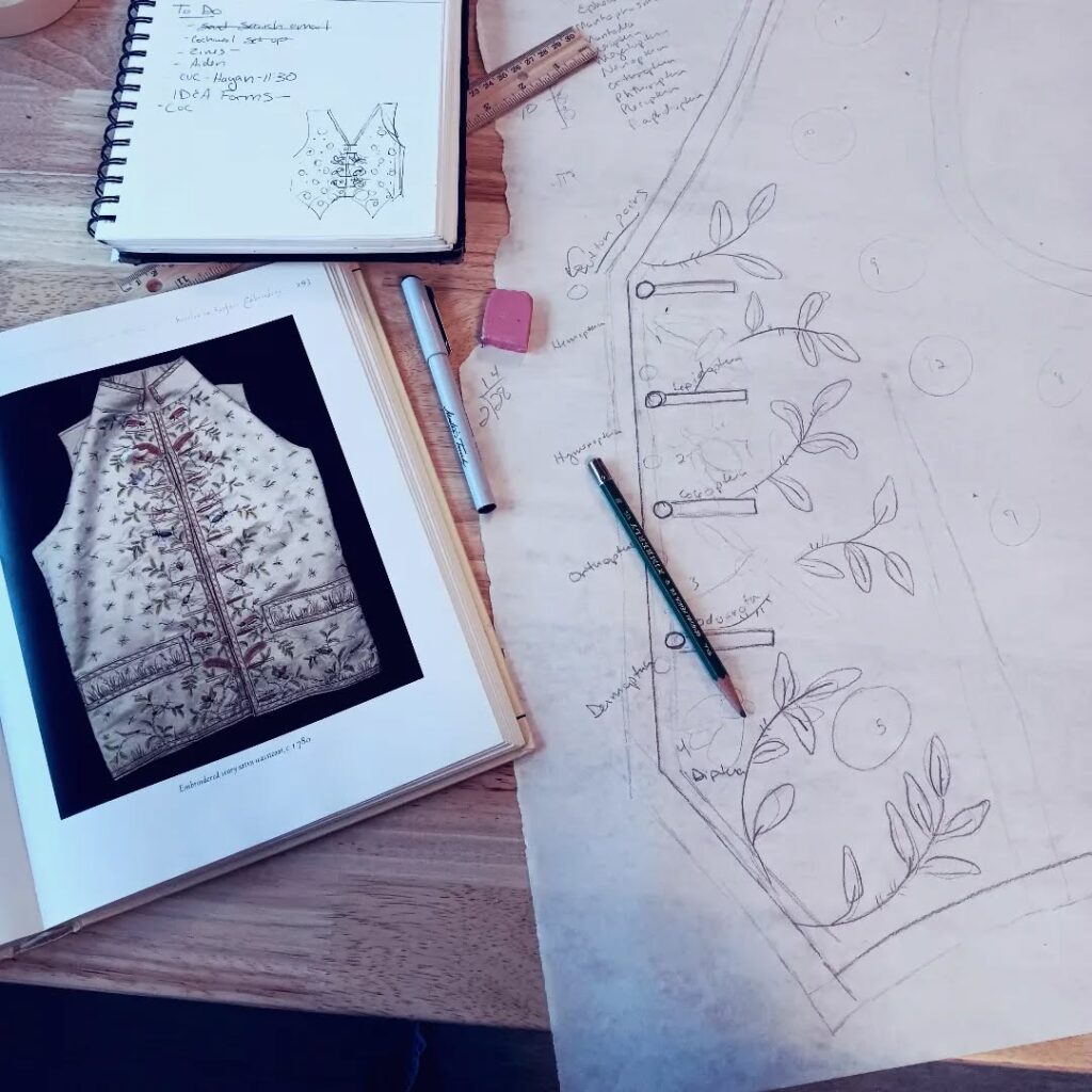 An open book with a page showing a vest-like garment. Next to it a sketch book and a sheet with a drawing of a vest pattern and other markings.