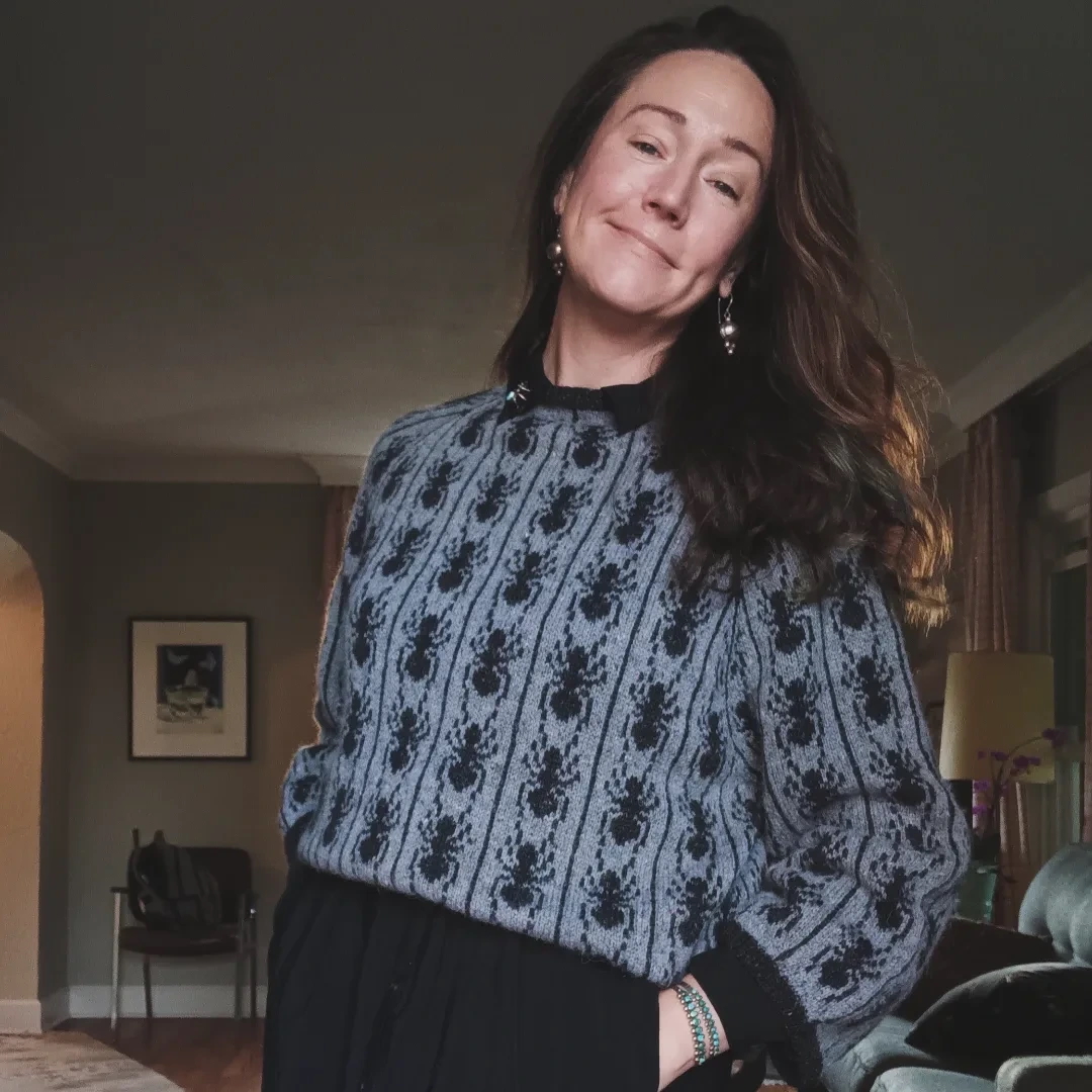 A person of light skin tone smiling. They have long hair and are wearing a knit sweater with an ants pattern.