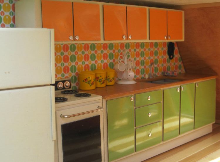 The inside of a miniature dollhouse featuring a kitchen with colorful cabinets, a stove and refrigerator