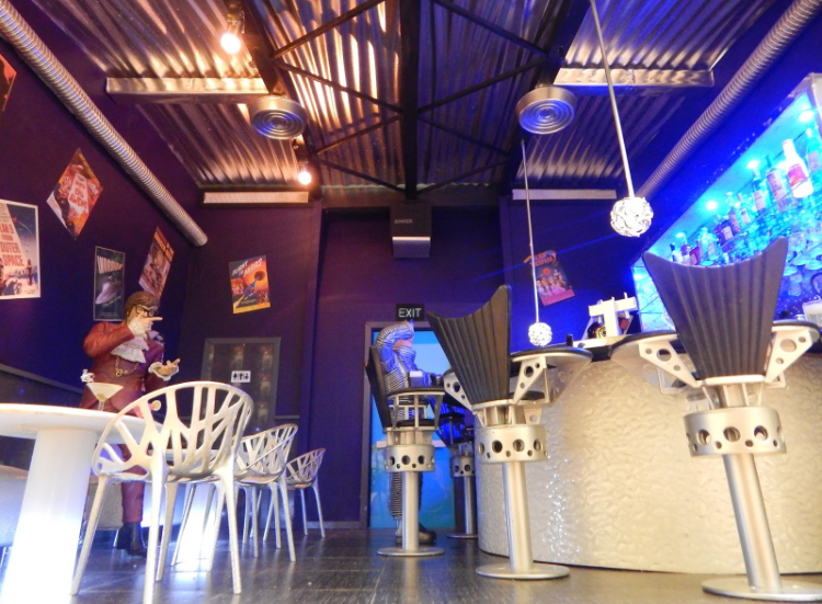 The inside of a miniature dollhouse featuring a bar-like space with high stools and other furniture