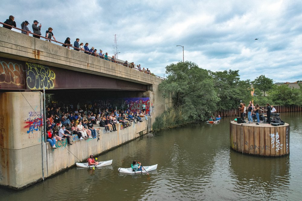 A wide shot capturing a bridge going over a river during the summertime, with people sitting and standing under the bridge and a band playing music atop a river structure surrounded by a few kayakers.