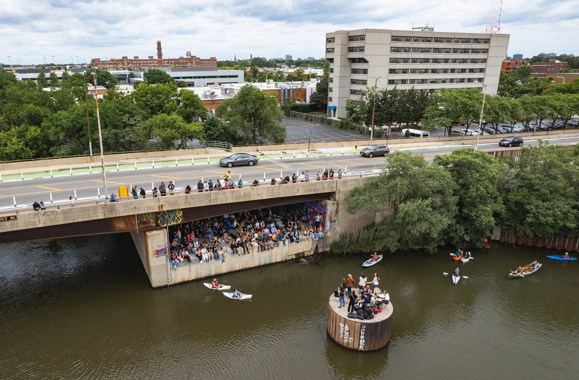 A wide shot capturing a bridge going over a river during the summertime, with people dotting the bridge and a band playing music atop a river structure surrounded by a few kayakers.