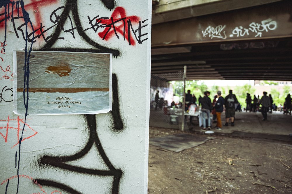 A placard with coordinates reads 'high noon' and a date on a structure with graffiti as a crowd gathers behind.