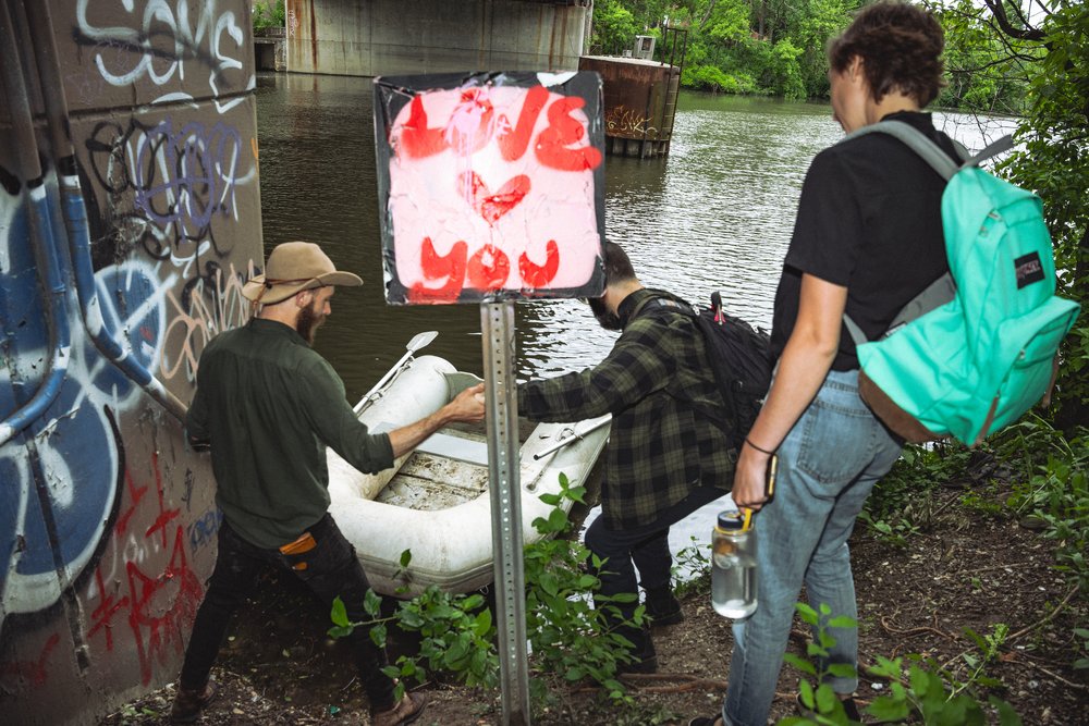 Three people with light skin stand before an inflatable boat near a body of water, behind a sign with red graffiti reading 'love you.'