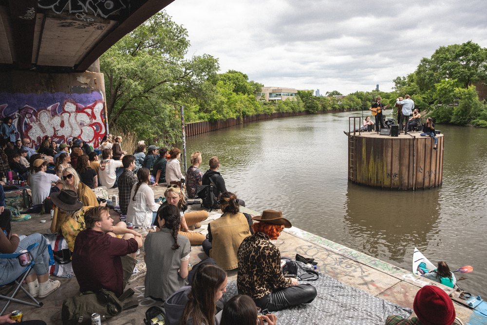 A crowd sits under a bridge underpass next to graffiti while overlooking a band playing on a floating river structure.