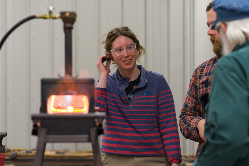 A light-skinned person in a striped sweater puts ear protection in her ear while wearing clear glasses and standing next to a hot forge.