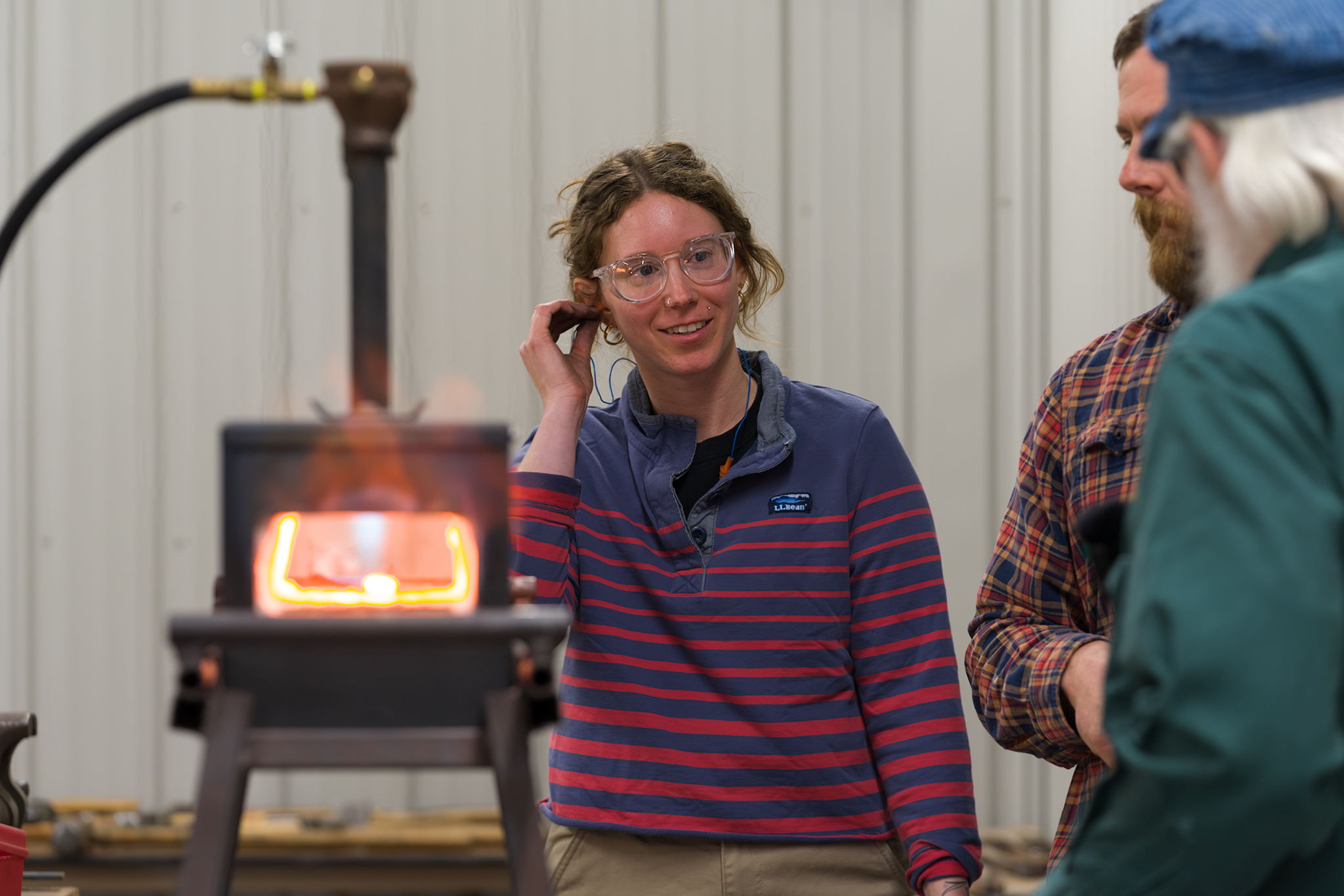 A light-skinned person in a striped sweater puts ear protection in her ear while wearing clear glasses and standing next to a hot forge.