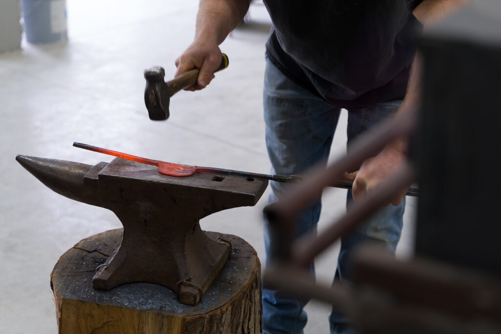 A hand holds a hammer while another hand holds metal that is hot and orange on the opposite side.