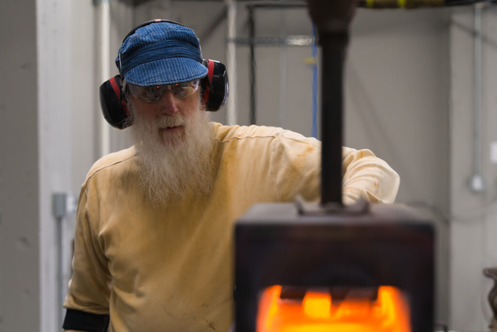 A light-skinned man in a blue hat wearing ear protection faces a forge with fire inside.