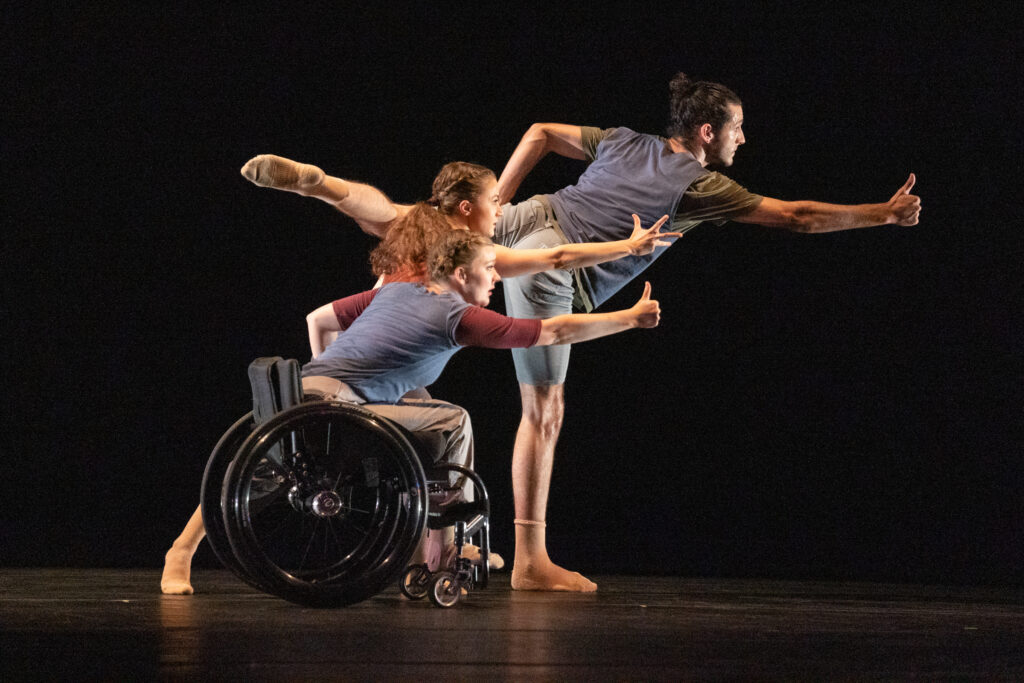 Three dancers wearing performance clothes in muted tones are in a line formation of decreasing height order from back to front. Each dancer is facing right and reaching their right arm out to make a thumbs-up gesture.