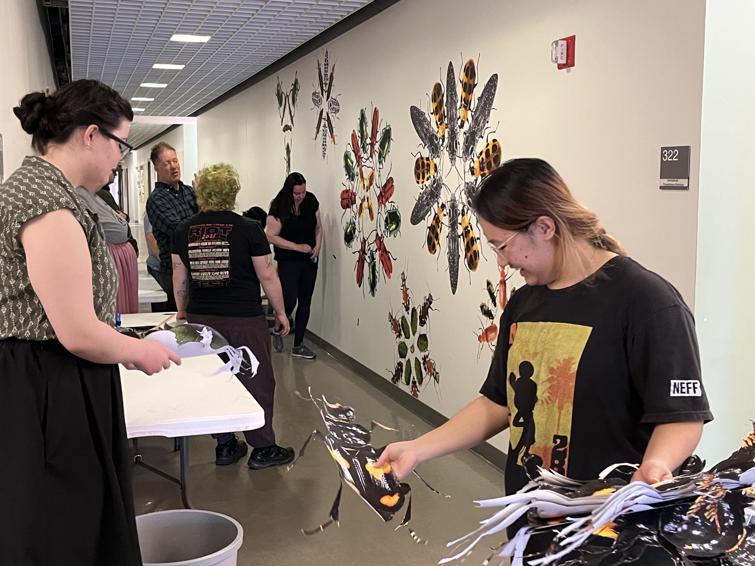 People standing by a hallway wall with an in-progress mural depicting beetles. Some people are holding cut-out photos of beetles that will go on the mural wall.