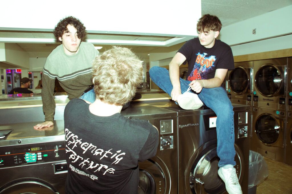 Two people sitting on laundromat washing machines while a third person stands in front of them.