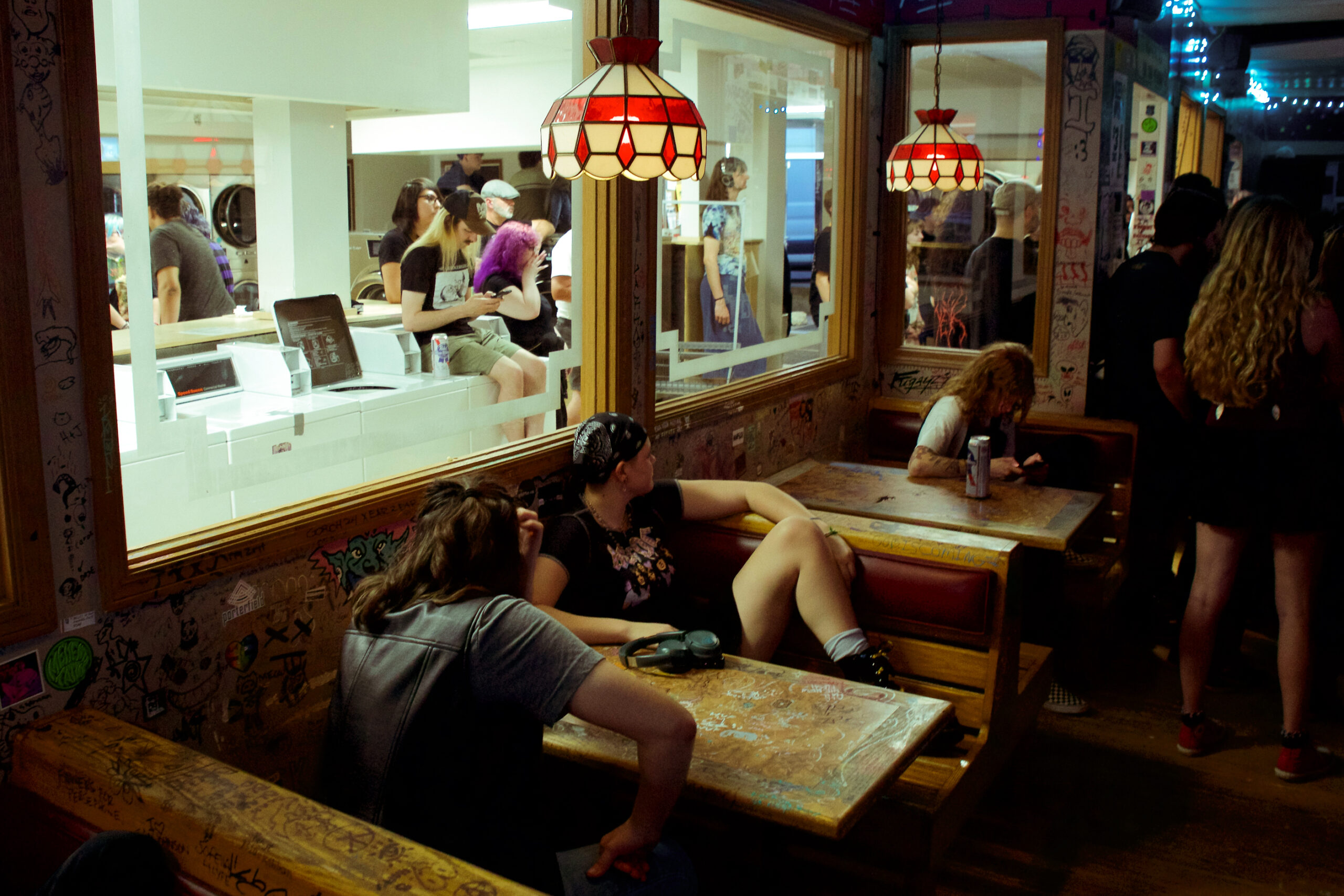People sitting at booths with marked up wooden tables and benches in a dimly lit room. Others are standing looking in one direction. In the background, through glass windows, is a laundromat with people standing around or sitting on laundry machines.