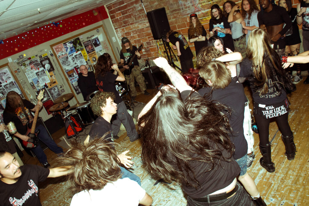 People standing around while some in the middle of the room are dancing or moshing. There's a band in the background playing instruments and a person singing into a microphone.