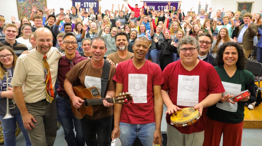 A group of musicians and students pose for a joyful group photo at UW-Stout. In the front row, musicians hold Brazilian percussion instruments and wear labeled shirts. Behind them, a large, smiling crowd of students cheers with hands raised.
