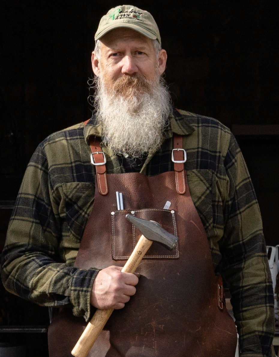 Light-skinned man wearing a baseball cap, flannel shirt, and apron, holding a metal and wooden hamer looks directly at the camera.